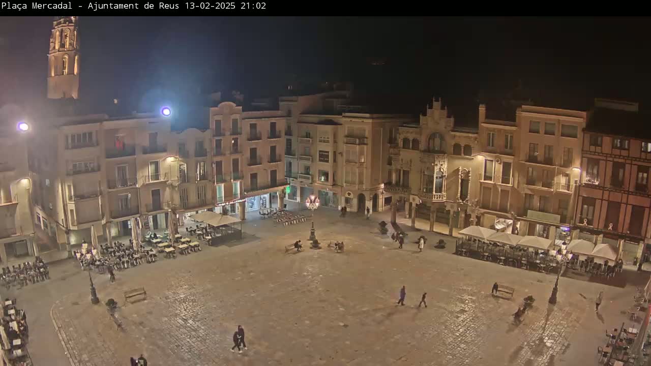A nighttime, high-angle view shows a mostly empty town square surrounded by illuminated buildings, with a few people walking around and some outdoor seating areas.