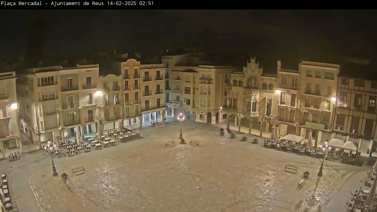 A mostly empty town square at night, illuminated by streetlights, with several outdoor cafes around its perimeter under mostly clear skies.