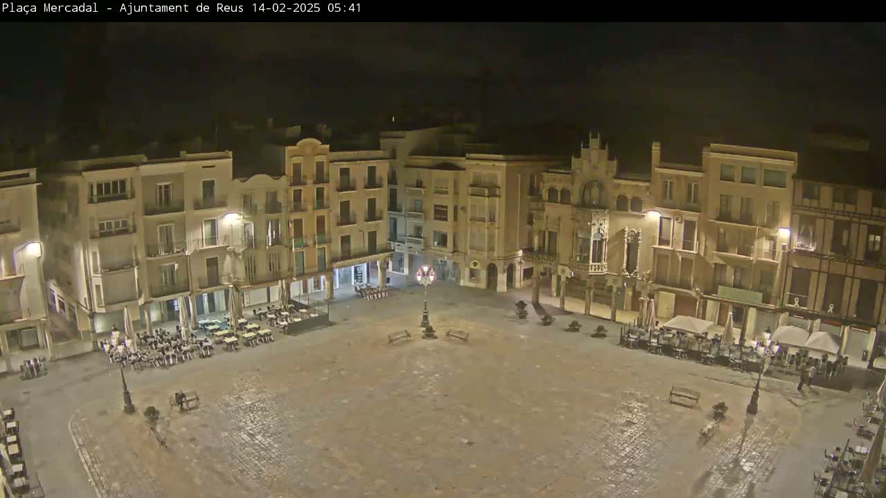 An empty, cobblestone town square at night, surrounded by multi-story buildings with outdoor seating areas at some of the ground-floor businesses.