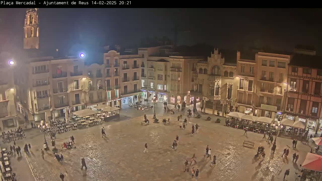 A nighttime, bird's-eye view of a town square with people walking around and several outdoor cafes, all illuminated under the dark sky.