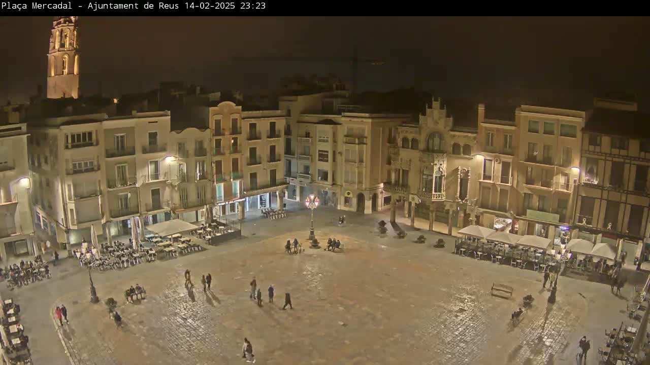 A nighttime view of a large, mostly empty town square surrounded by multi-story buildings, with a few people and outdoor seating areas visible.