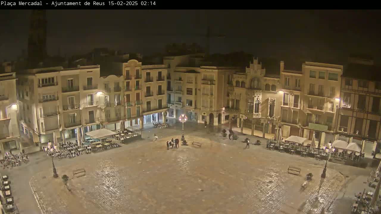 A wet, empty town square at night, surrounded by multi-story buildings with outdoor seating areas.