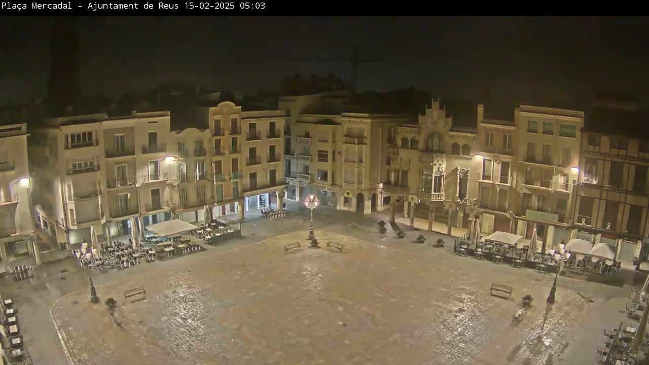 A rain-slicked, empty town square at night is surrounded by multi-story buildings with lights illuminating the scene.