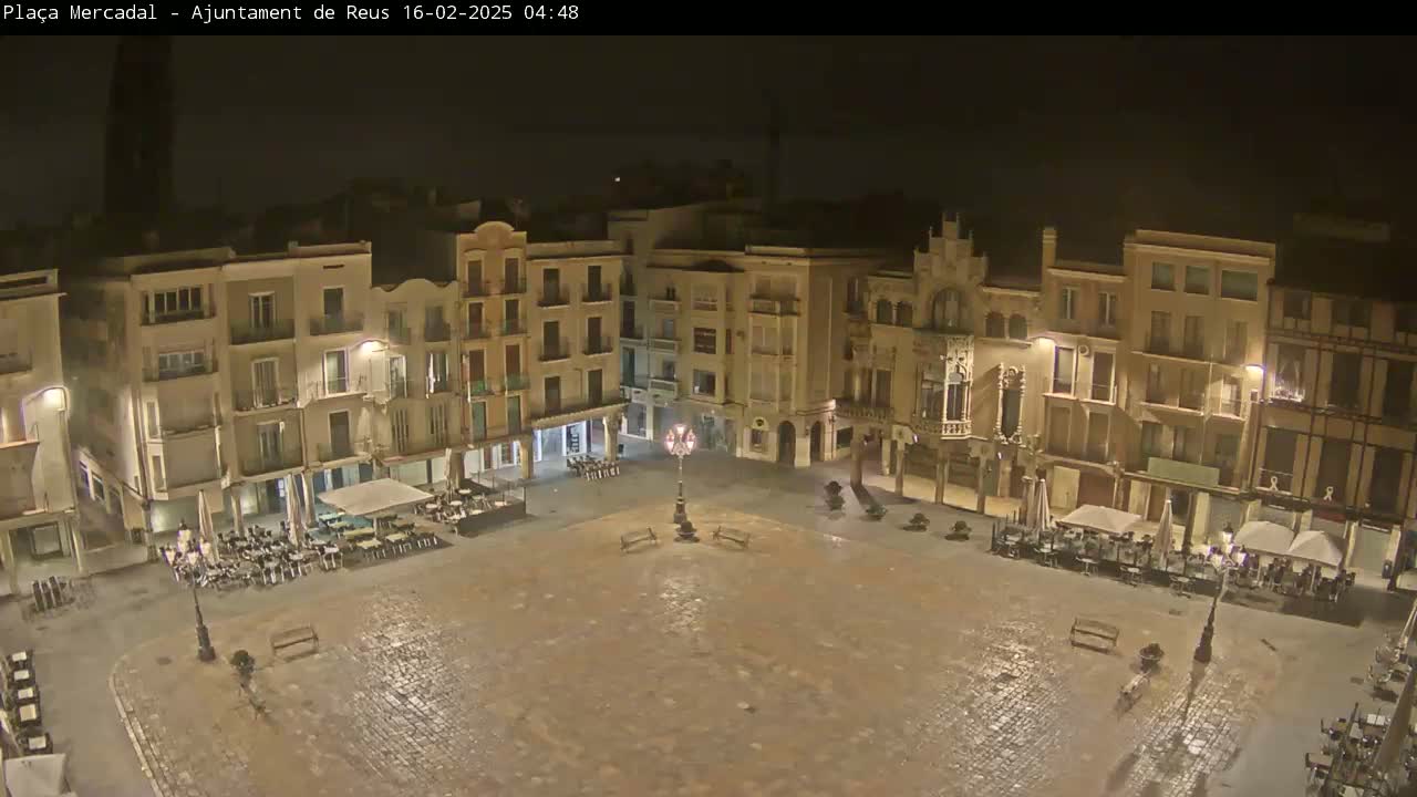 An empty, cobblestone town square at night, surrounded by buildings with illuminated windows and outdoor seating areas, under a dark sky.