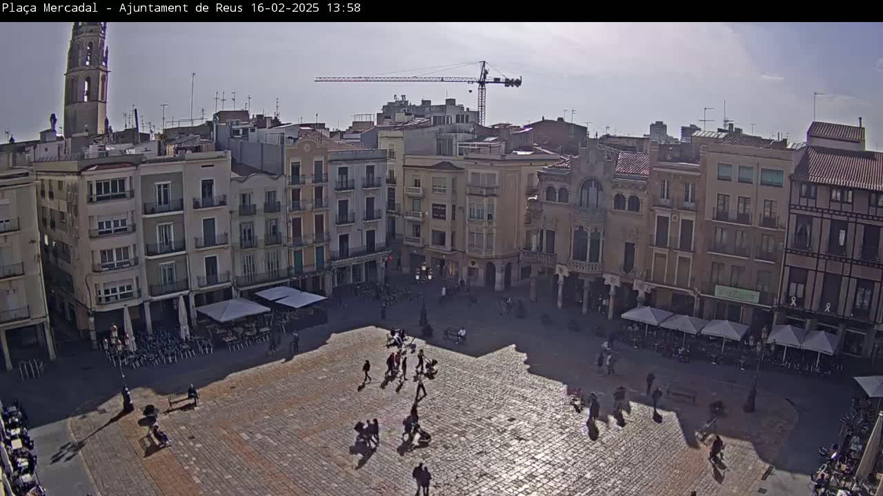 A sunny day reveals a large, cobblestone town square surrounded by multi-story buildings, with people sparsely scattered throughout and a few outdoor cafes set up.