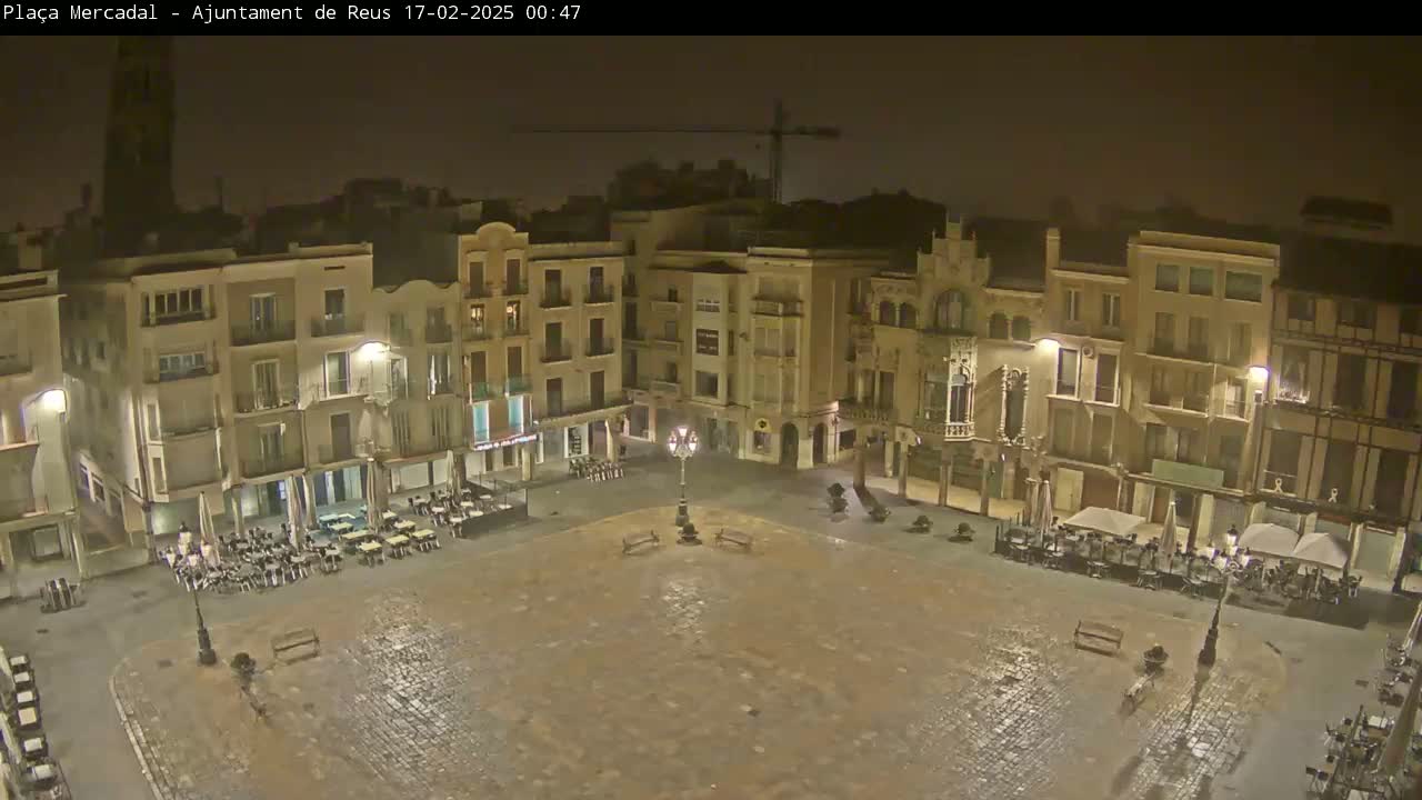 A wet, empty town square at night, surrounded by multi-story buildings with illuminated windows and sparsely occupied outdoor seating areas.