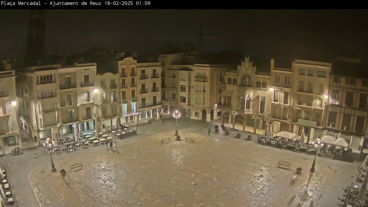 A mostly empty, cobblestone town square at night, surrounded by multi-story buildings with outdoor seating areas at some of the ground-floor businesses.