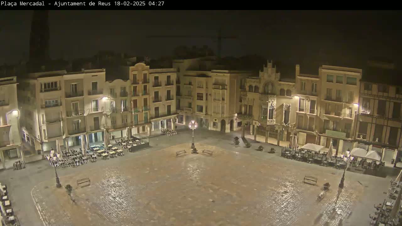 An empty, cobblestone town square at night, surrounded by multi-story buildings with outdoor seating areas, is lit by streetlights.