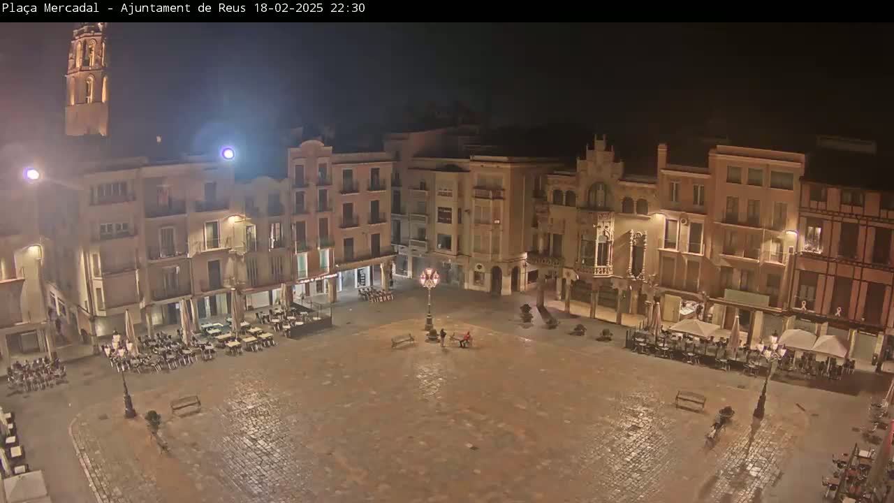A nighttime view of a town square with several buildings surrounding it, some with outdoor seating, under a dark sky.