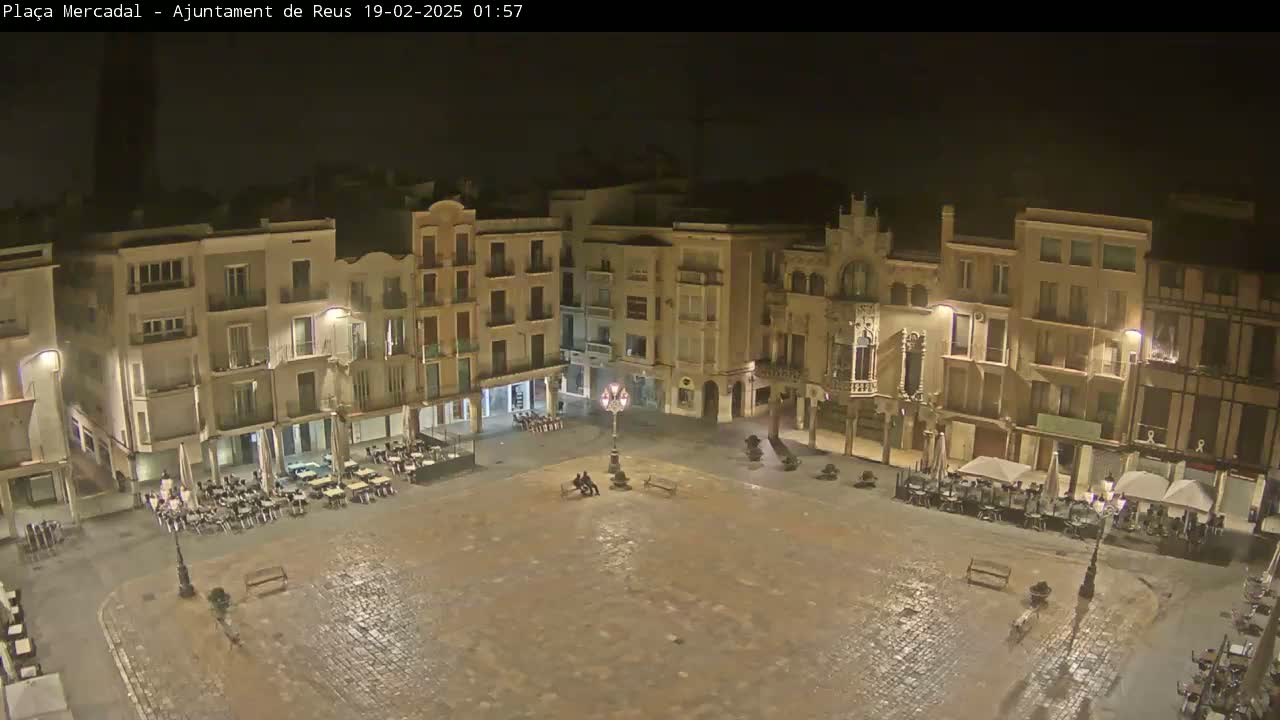 An empty, cobblestone town square at night, surrounded by multi-story buildings with outdoor seating areas at ground level, is lit by streetlights.