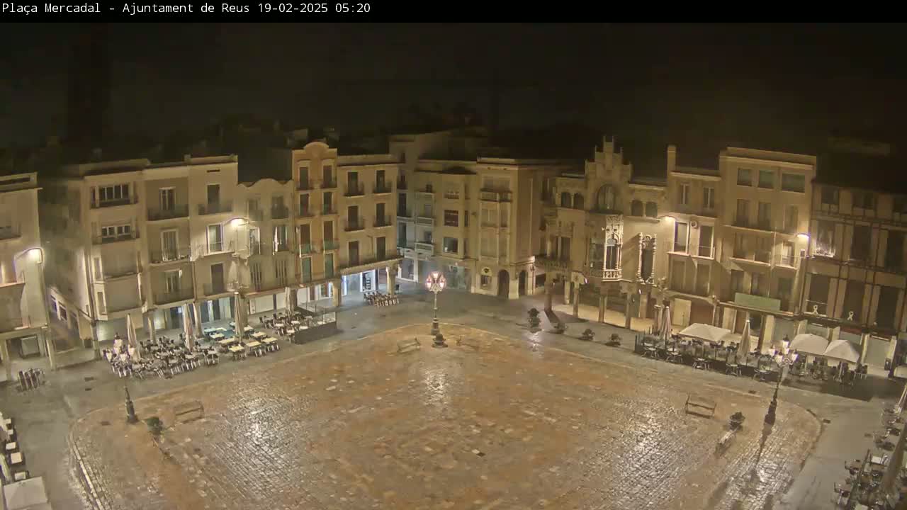 A rain-slicked, cobblestone town square at night, surrounded by illuminated buildings with outdoor seating areas.
