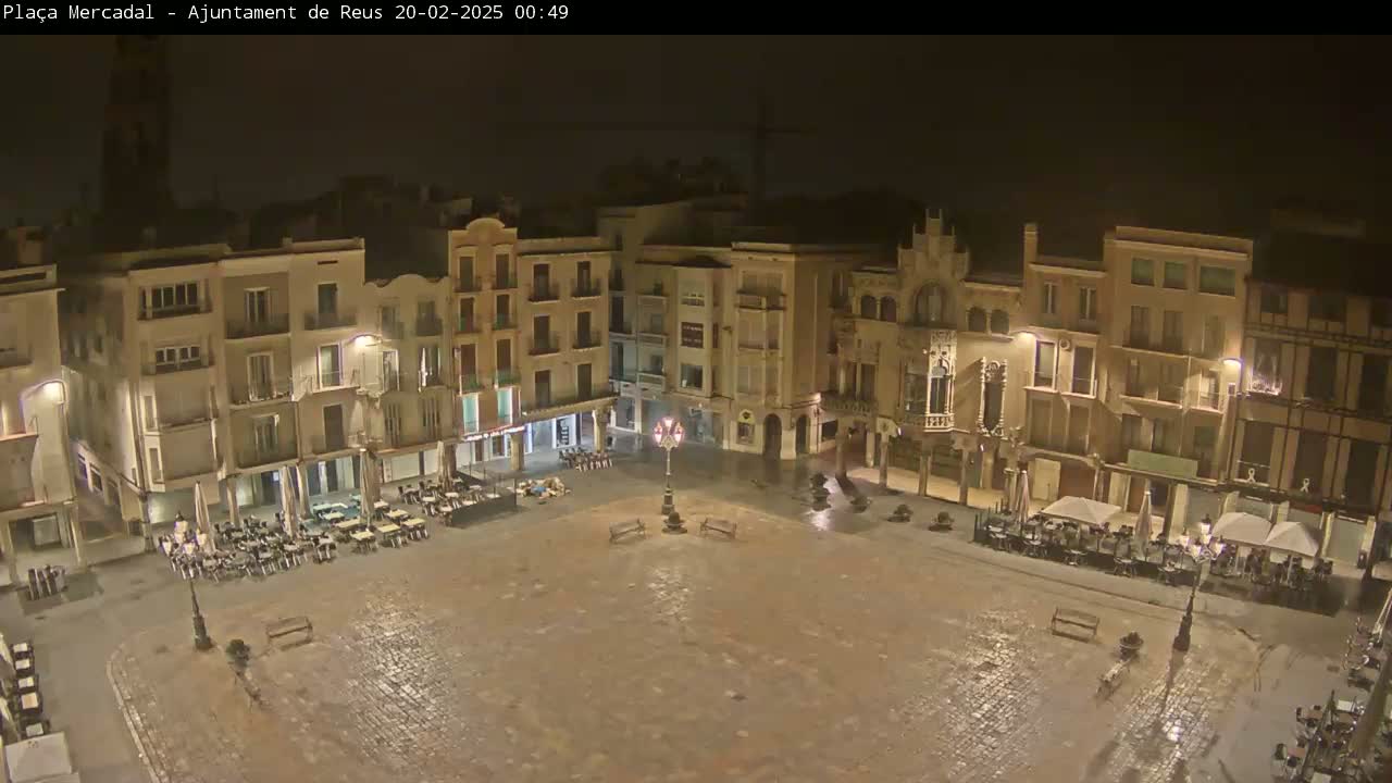 A wet, empty town square at night is surrounded by multi-story buildings with outdoor seating areas.