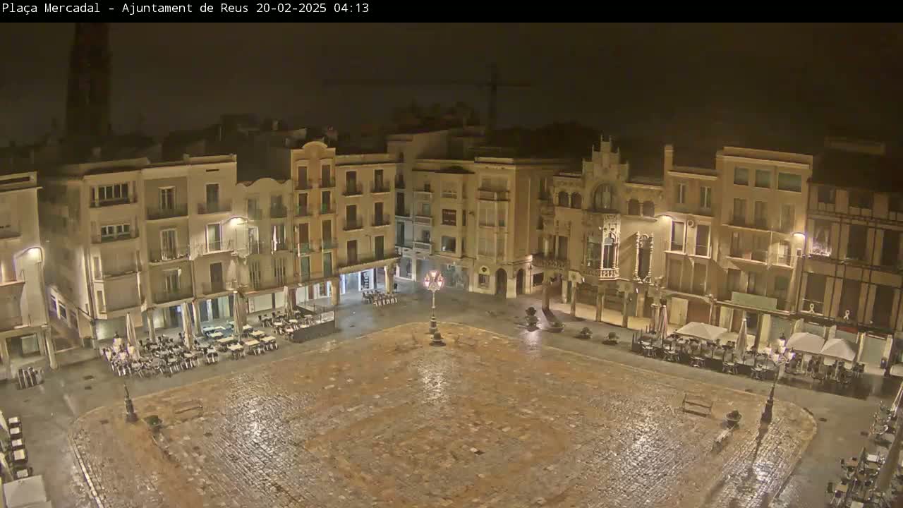 A rain-slicked town square at night, surrounded by illuminated buildings with outdoor seating areas.
