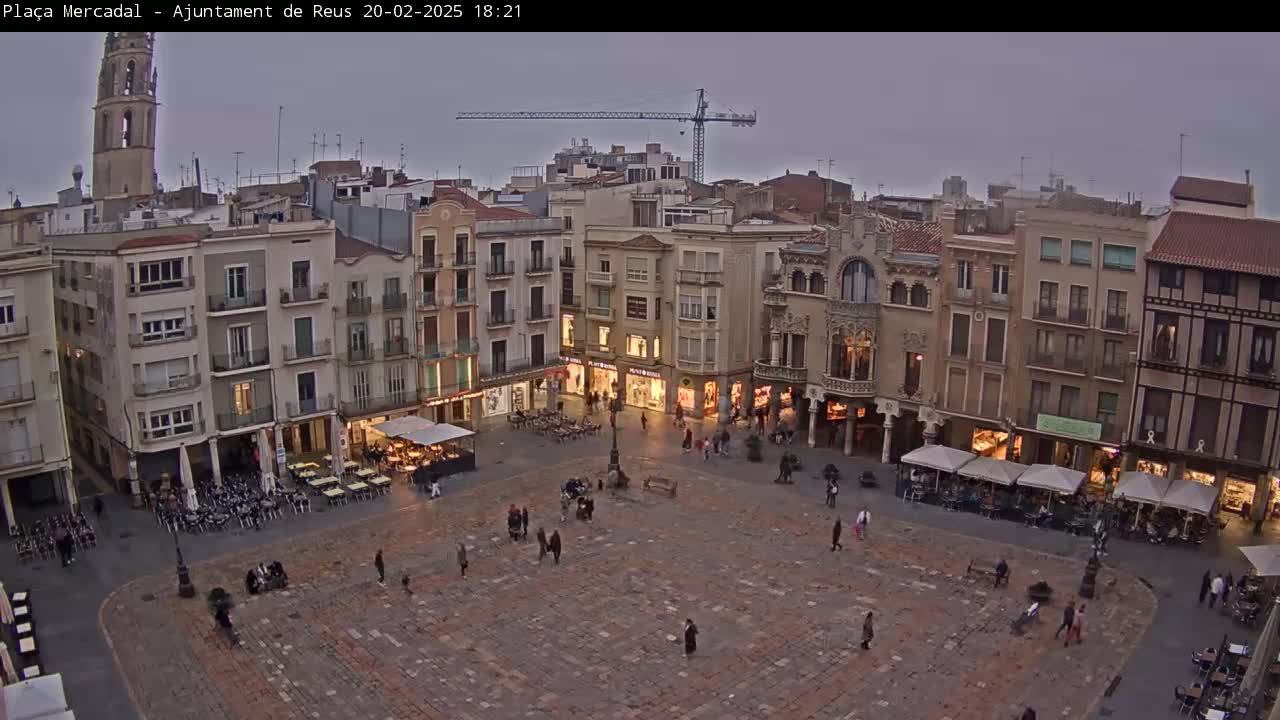 A large, stone plaza in a city is populated by a few people and outdoor cafes under an overcast sky.