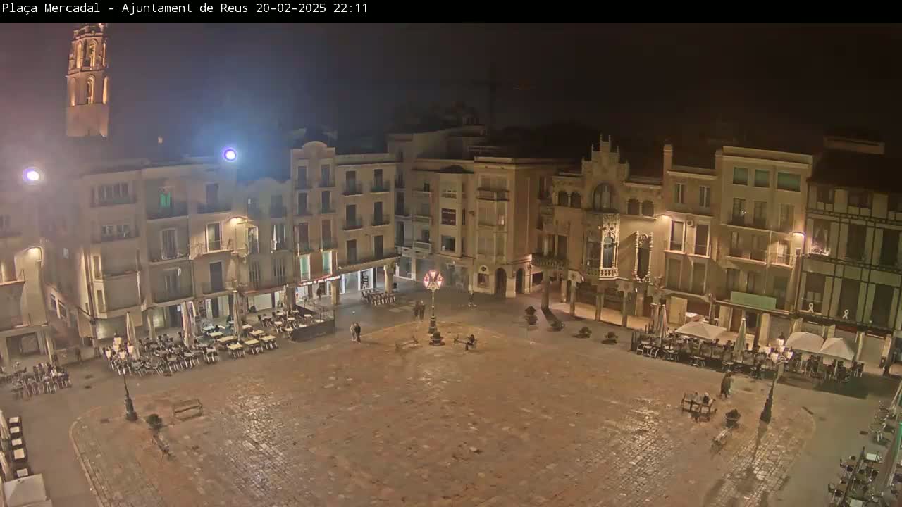 A mostly empty cobblestone town square at night, surrounded by multi-story buildings with outdoor seating areas at ground level, is lit by street lamps and building lights.