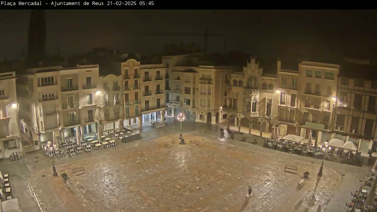 A rain-slicked town square at night, surrounded by multi-story buildings with outdoor seating areas.