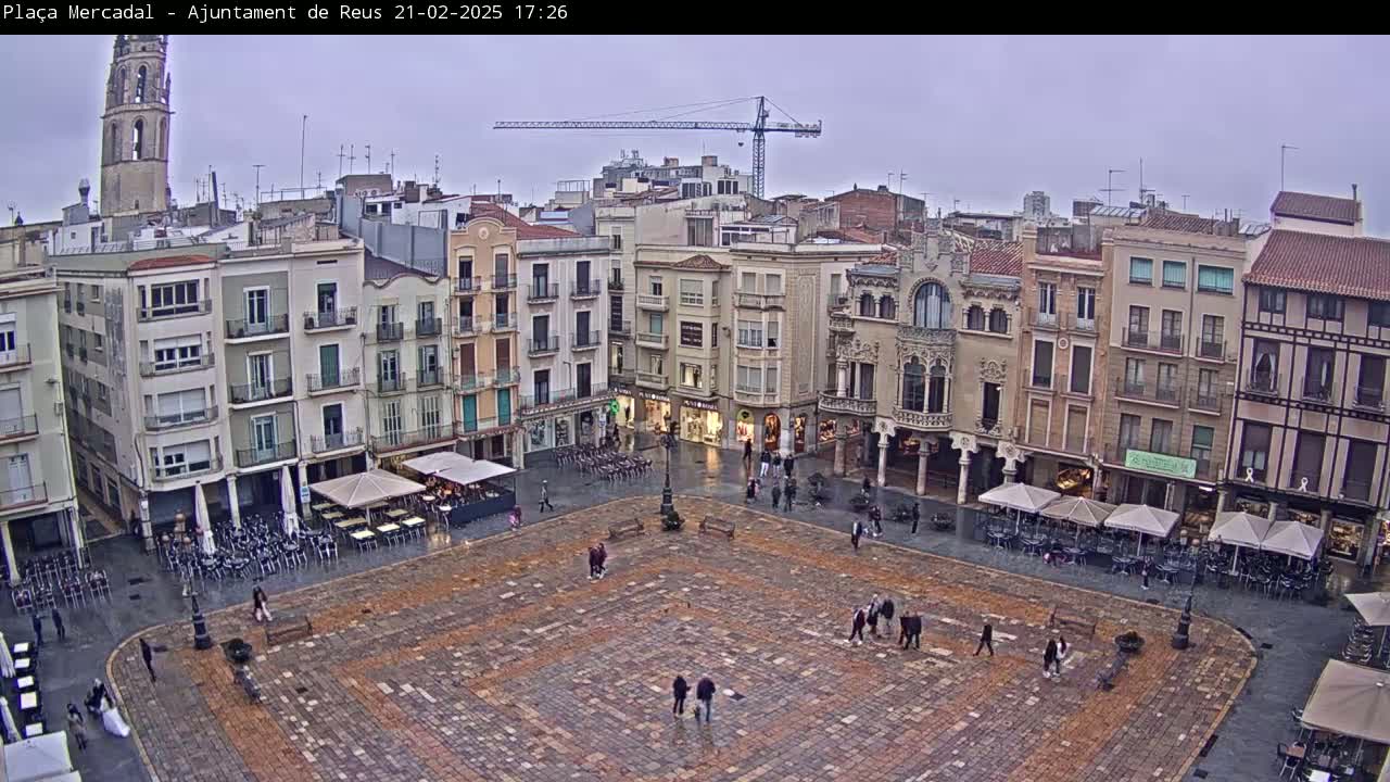 A large, paved town square, surrounded by multi-story buildings, is lightly populated with people under an overcast sky.