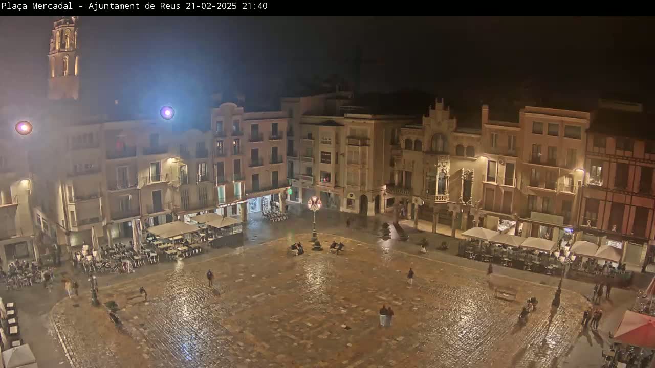 A rain-slicked town square at night, surrounded by illuminated buildings, with a few people scattered throughout.
