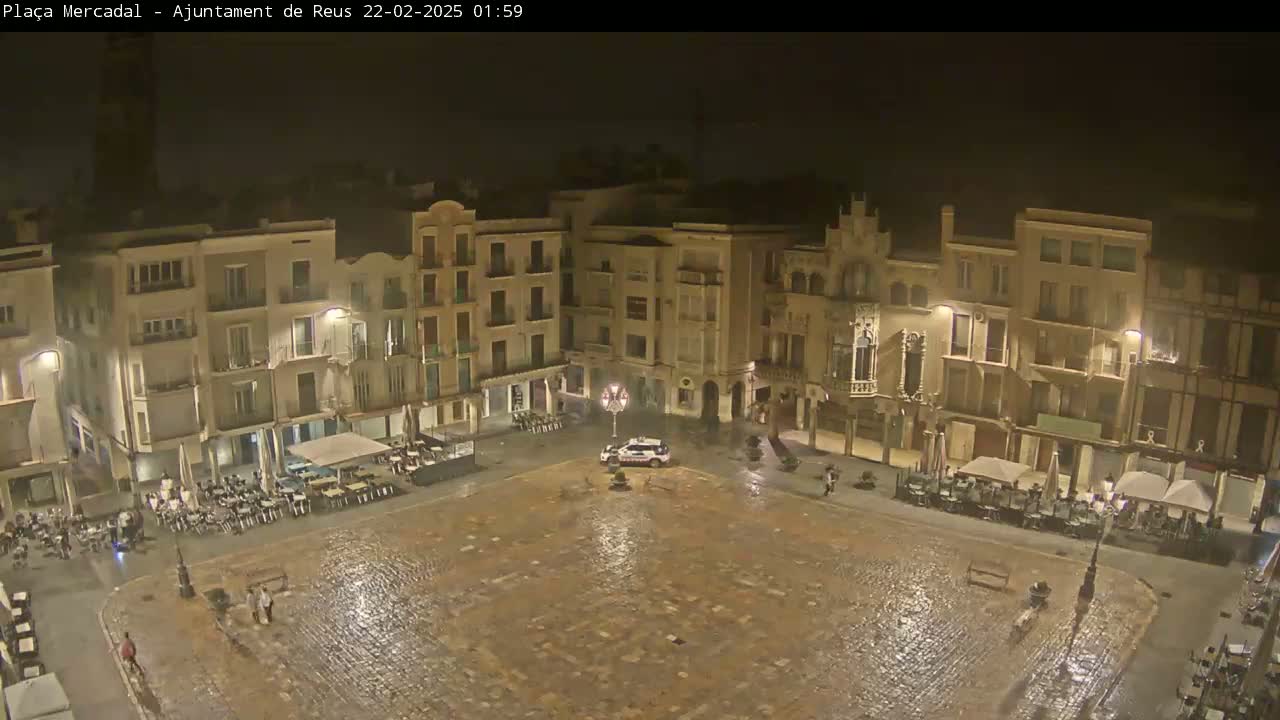 A rain-slicked, cobblestone town square at night, surrounded by multi-story buildings with outdoor seating areas, and a police car parked in the center.
