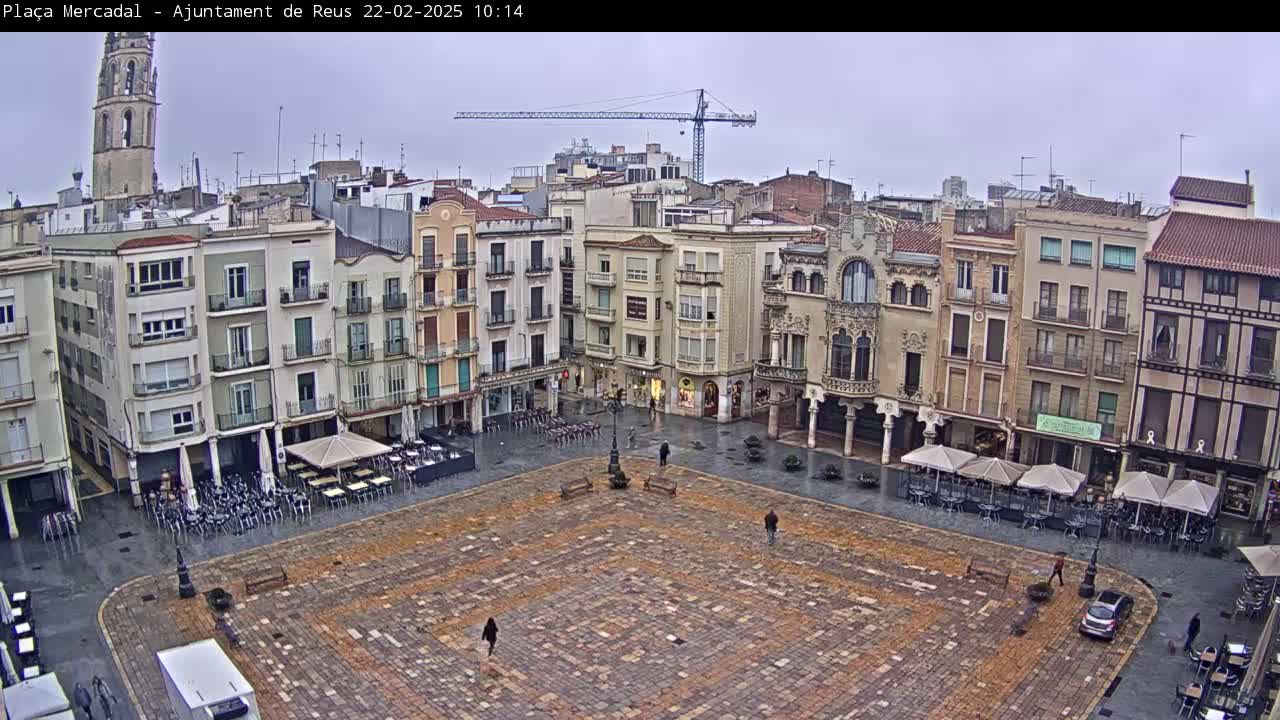 A large, mostly empty cobblestone town square is surrounded by multi-story buildings under an overcast sky; some outdoor café seating is visible.