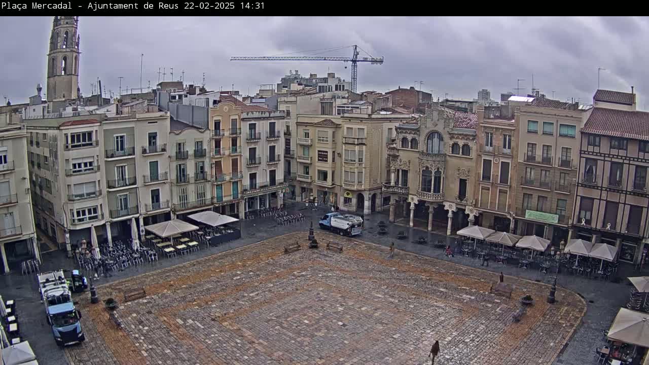 A large, cobblestone town square is surrounded by multi-story buildings under an overcast sky.