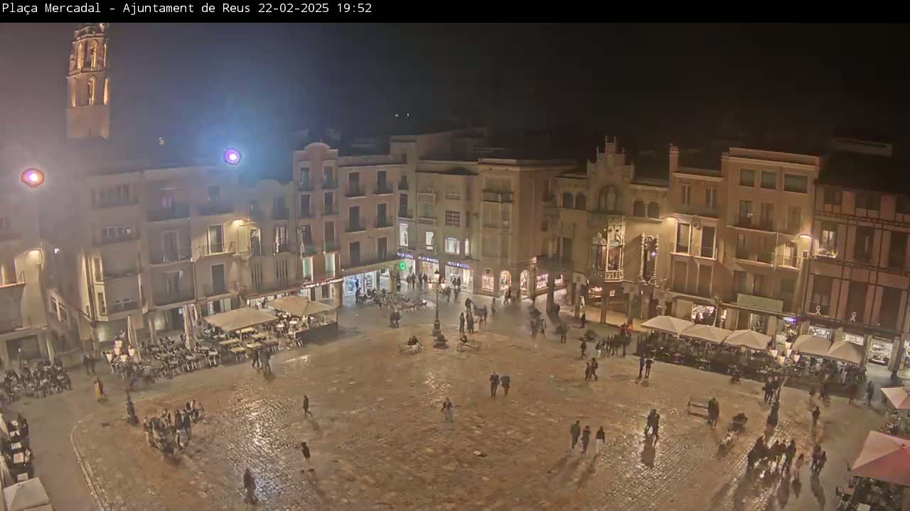 A town square at night, illuminated by streetlights, shows people walking around and sitting at outdoor cafes, with some wet pavement indicating recent rain.