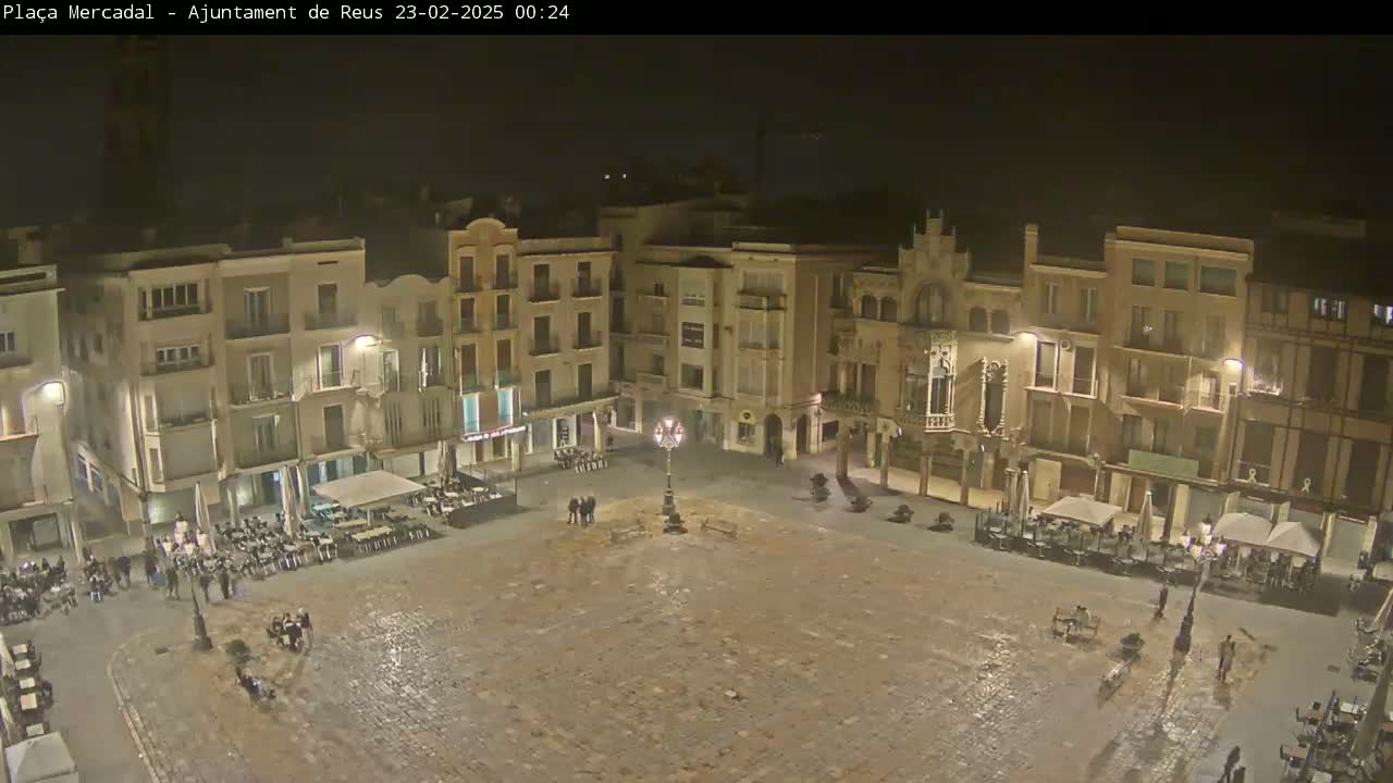 A mostly empty, cobblestone town square at night, surrounded by multi-story buildings with outdoor seating areas.
