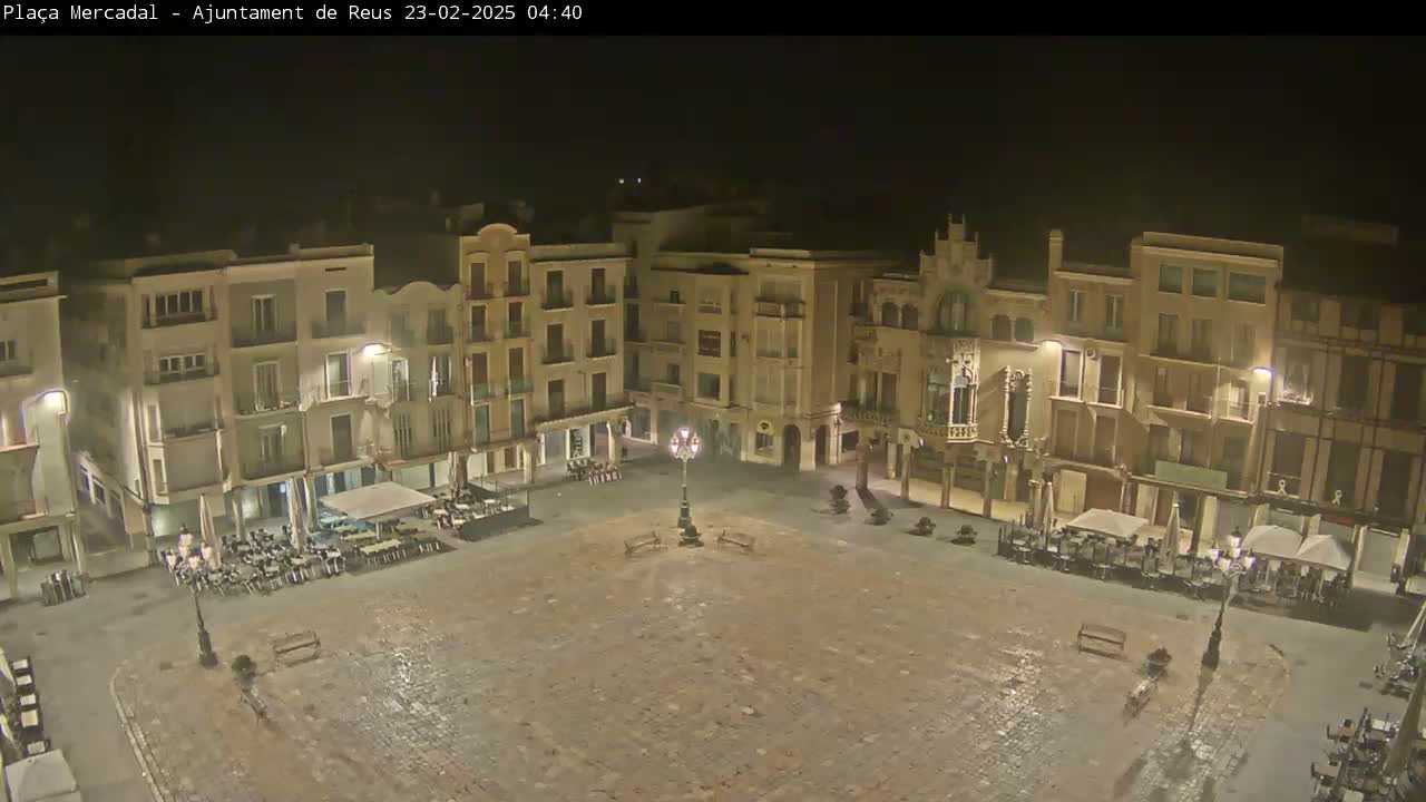 A mostly empty, cobblestone town square at night, surrounded by multi-story buildings with outdoor seating areas at some of the ground-floor businesses.