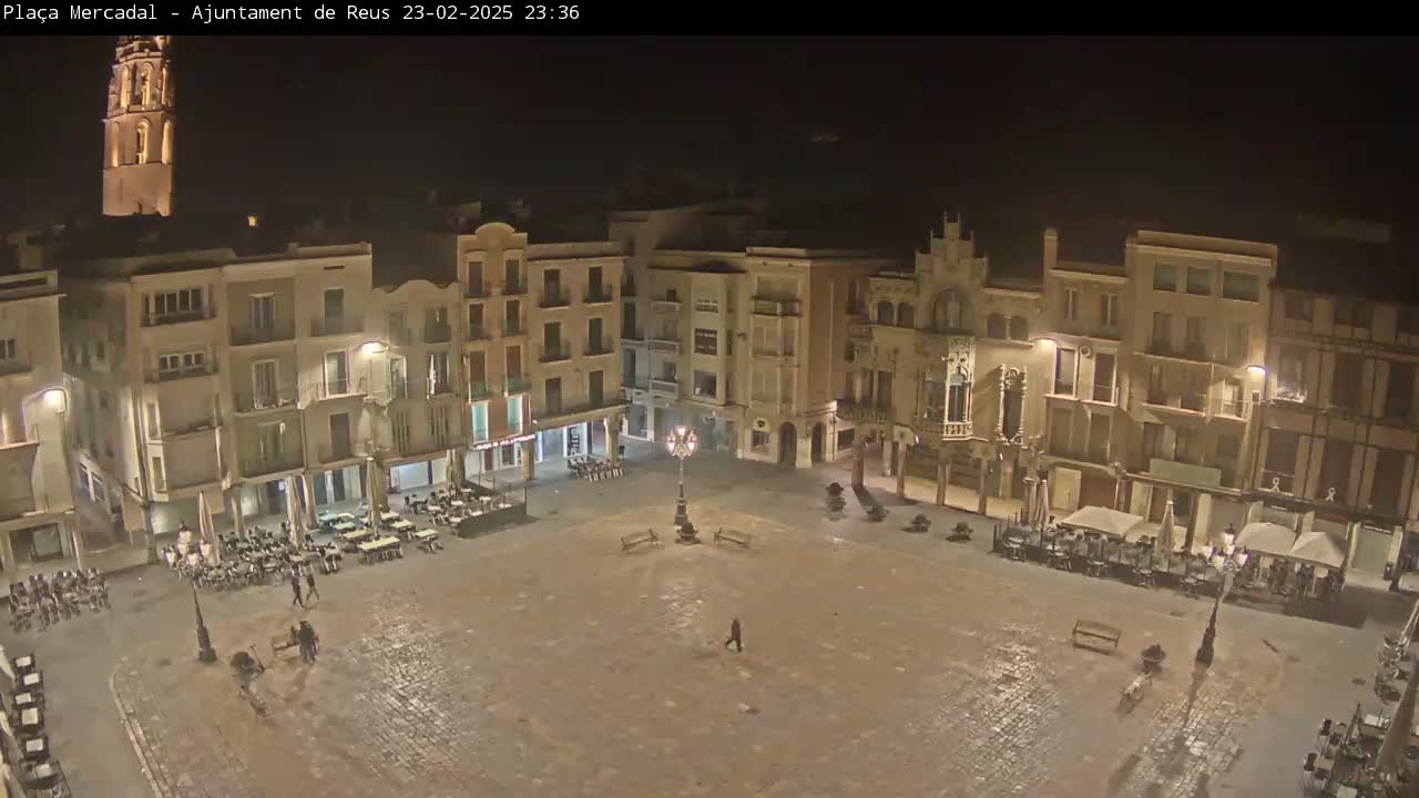 A mostly empty town square at night is surrounded by multi-story buildings with outdoor seating areas, and a tall tower is visible in the background.