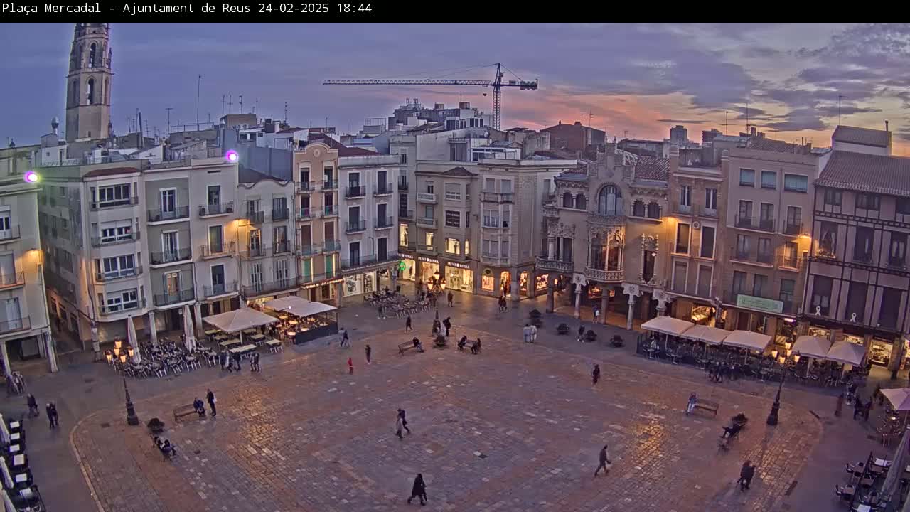 A large town square at dusk, under a partly cloudy sky, is surrounded by multi-story buildings with outdoor seating areas in front of some of the shops.