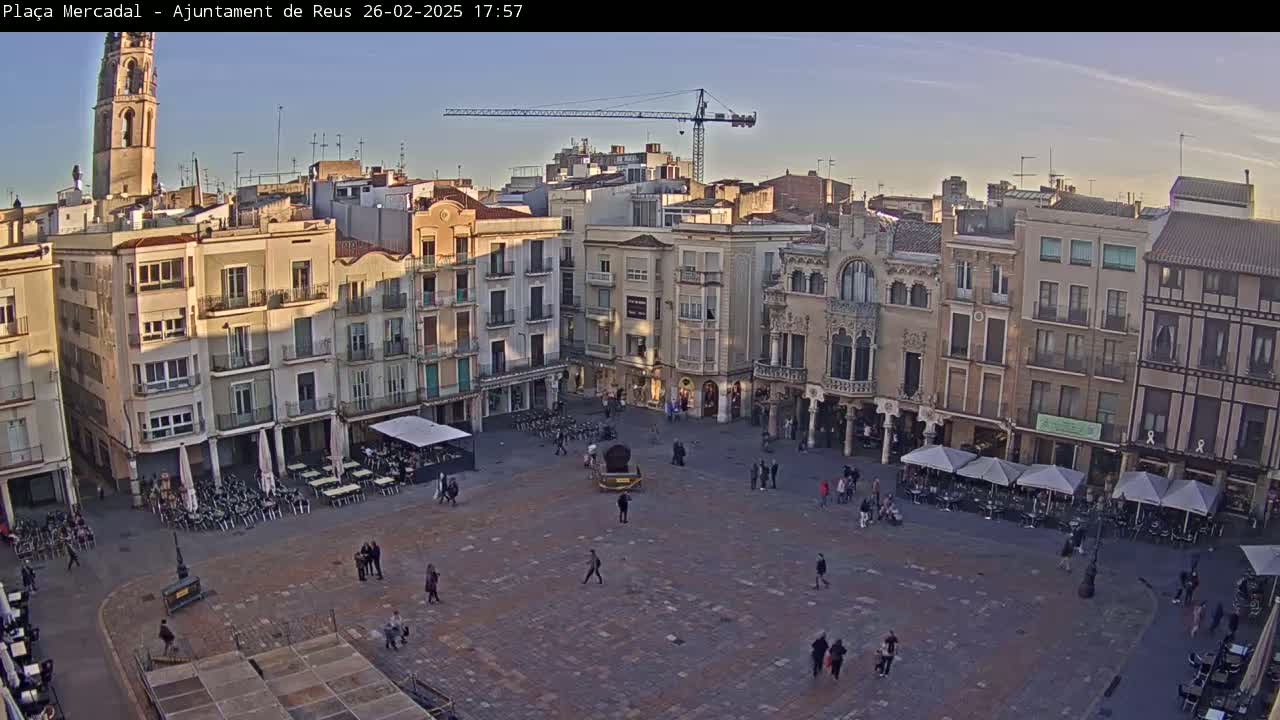 A large, paved town square is surrounded by multi-story buildings under a clear, twilight sky, with a few people walking around and some outdoor seating areas.