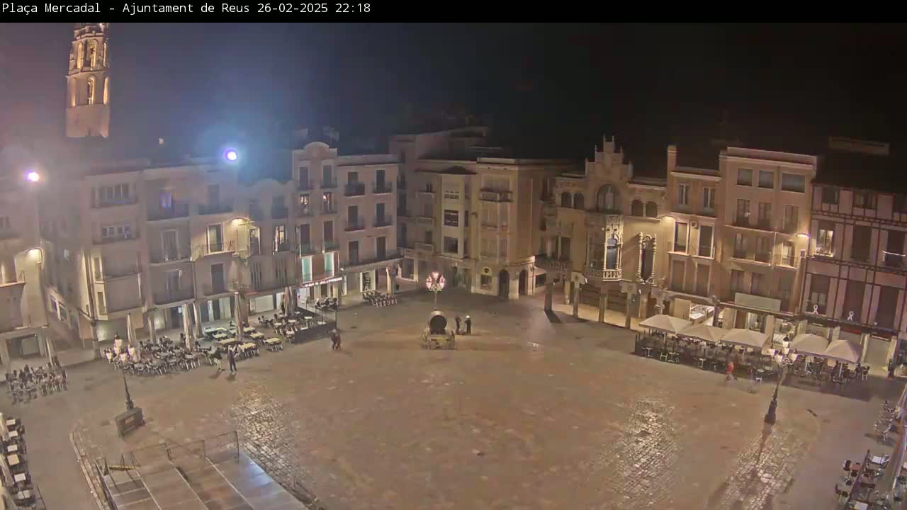 A nighttime, wet view of a town square surrounded by buildings with lights on, some with outdoor seating areas.