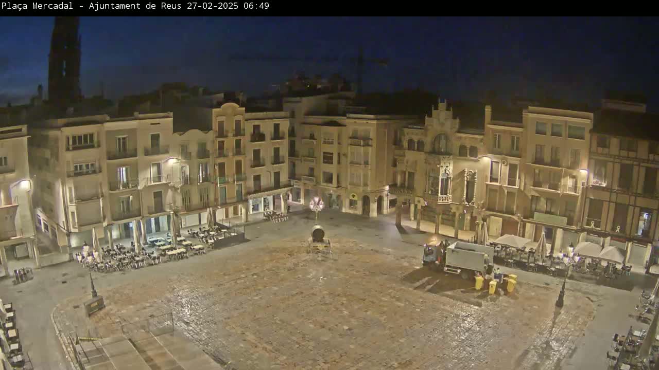 An empty town square at night, under a dark sky, is surrounded by buildings with outdoor seating areas.