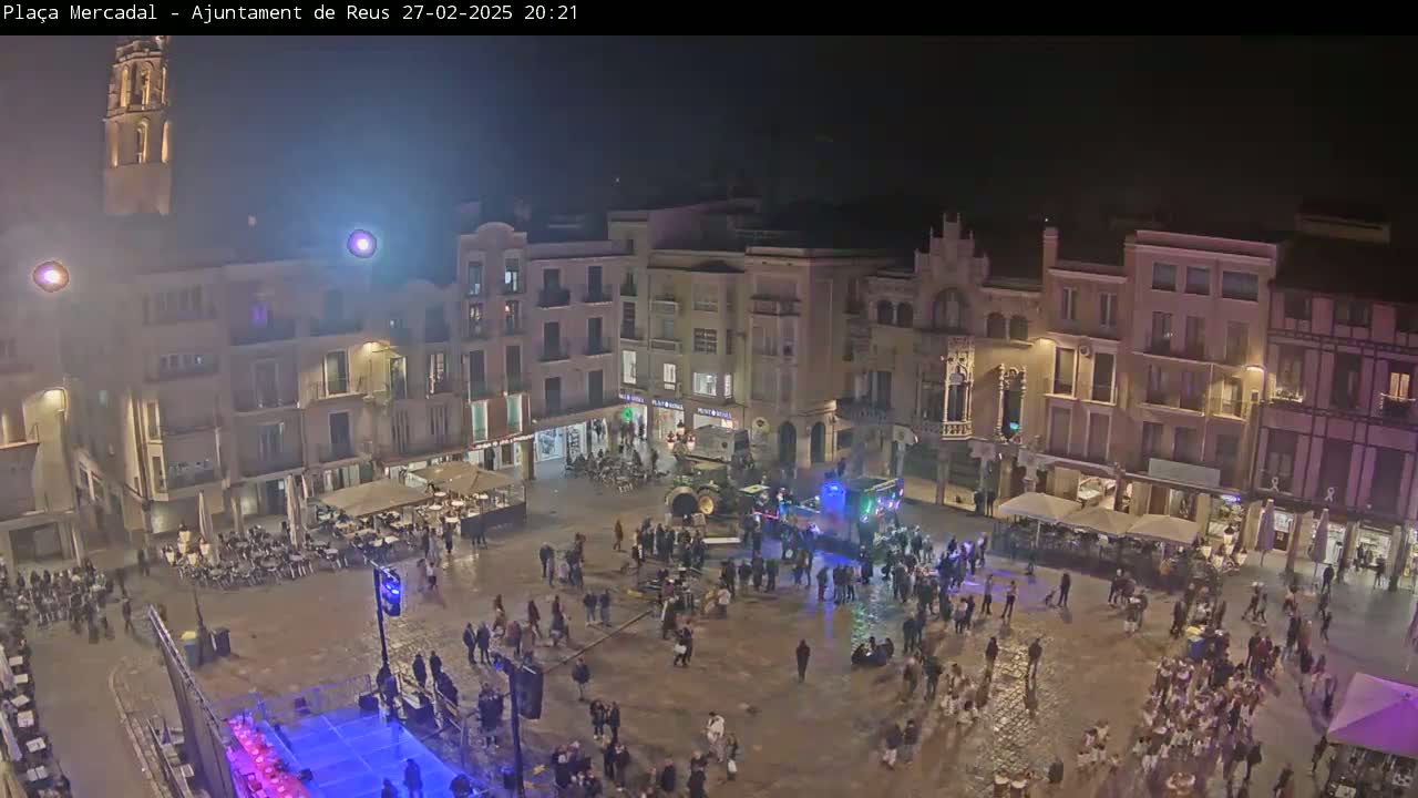 A nighttime, wet town square is filled with people gathered around a stage and several outdoor cafes under a dark sky.
