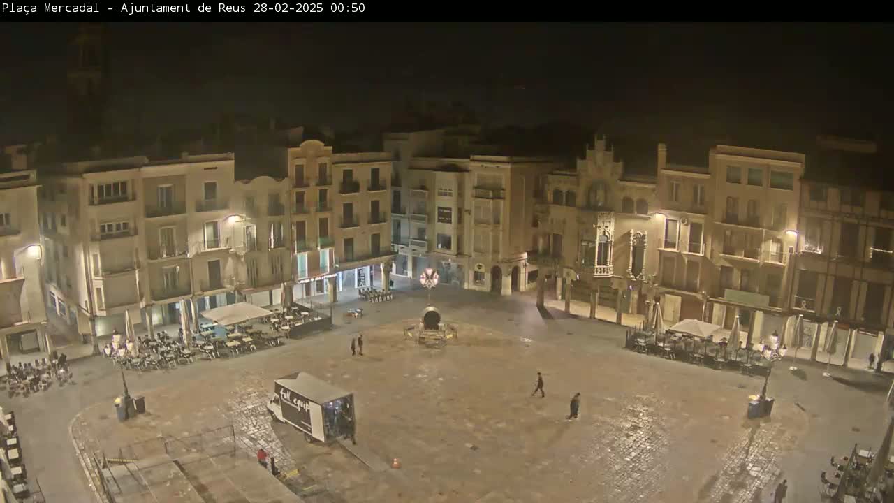 A mostly empty town square at night, surrounded by multi-story buildings with lights on, is slightly wet.