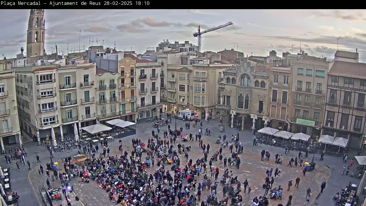 A large town square, under an overcast sky, is filled with people gathered in a crowd and surrounded by multi-story buildings.