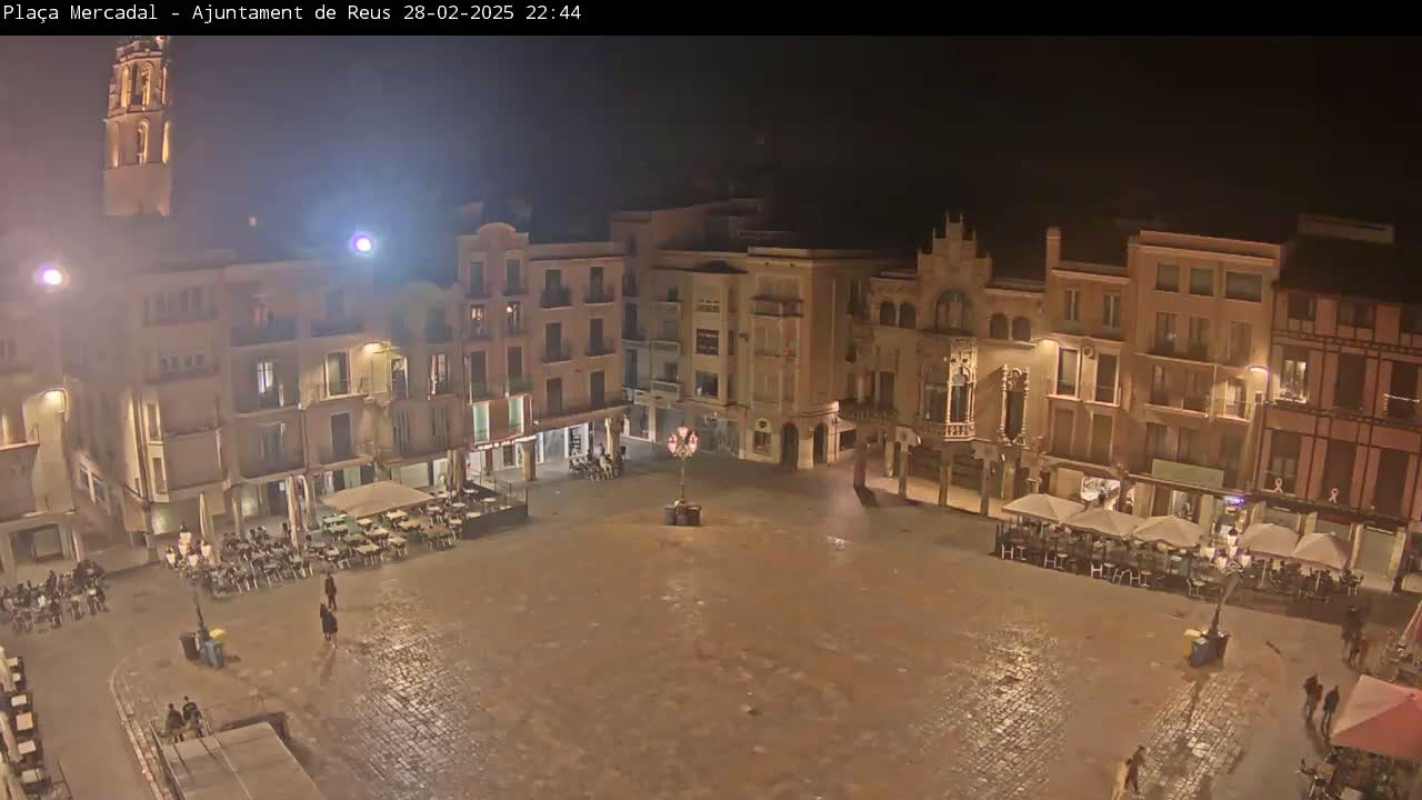 A mostly empty town square at night, illuminated by streetlights, is surrounded by buildings, some with outdoor seating areas.