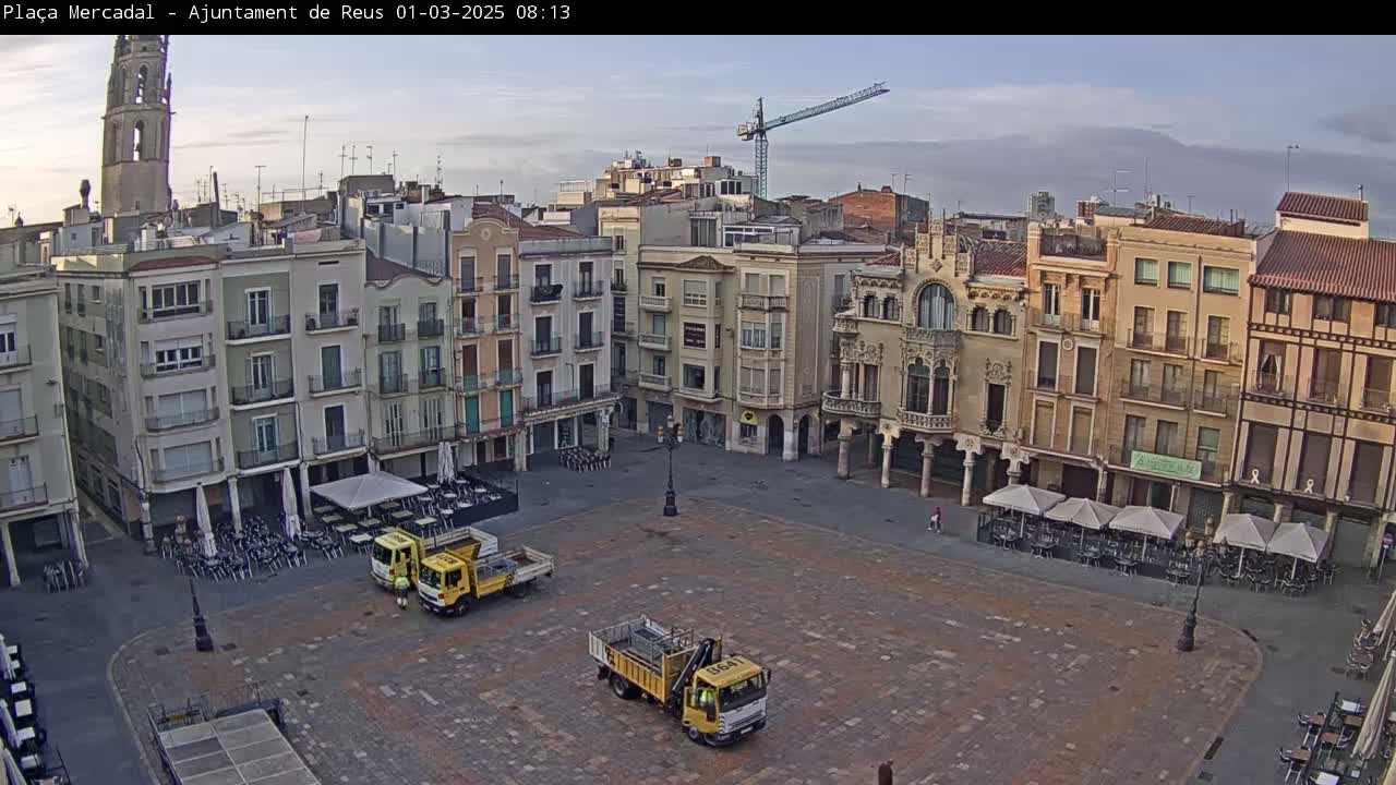 A mostly empty town square under a partly cloudy sky, with several buildings surrounding it, including a tall church tower, and a few yellow trucks parked in the square.
