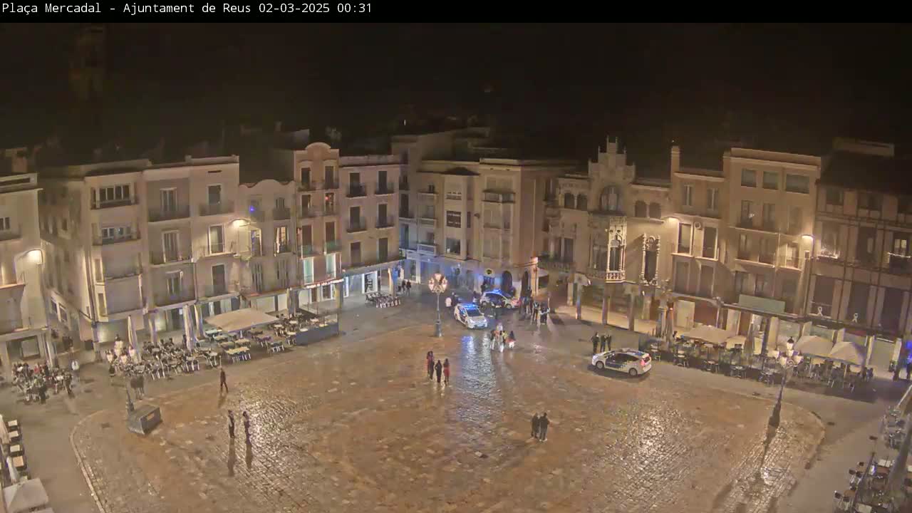 A wet, nighttime town square is surrounded by illuminated buildings, with several people and police cars present.