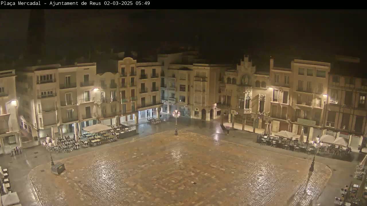 A rain-slicked town square at night, surrounded by multi-story buildings with outdoor seating areas.
