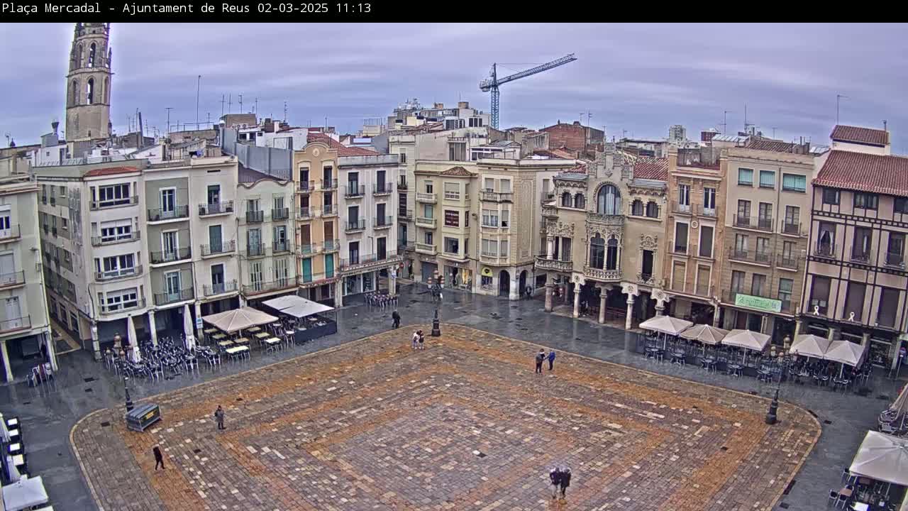 A large, mostly empty town square with multi-story buildings surrounding it under an overcast sky, with some people and outdoor cafe umbrellas visible.