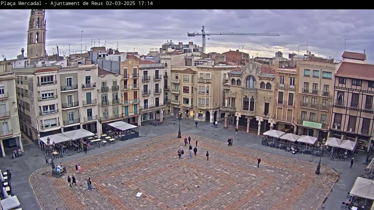 A large, paved town square is surrounded by multi-story buildings under a cloudy sky, with several people and outdoor cafe seating visible.