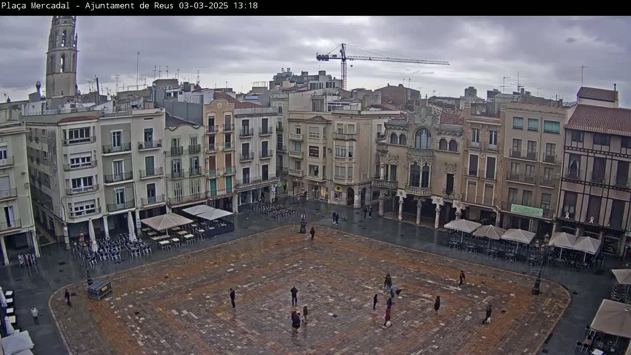 A large, paved town square with sparsely scattered people and outdoor cafe seating under umbrellas, surrounded by multi-story buildings under an overcast sky.