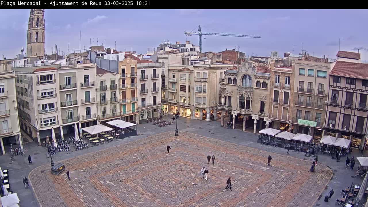 A large, cobblestone town square with several multi-story buildings surrounding it, under a mostly clear, dusky sky.