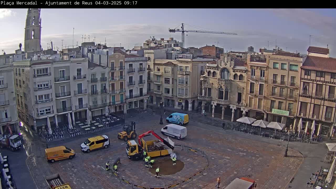A town square under a partly cloudy sky is being worked on by construction crews, with several vehicles and workers present.