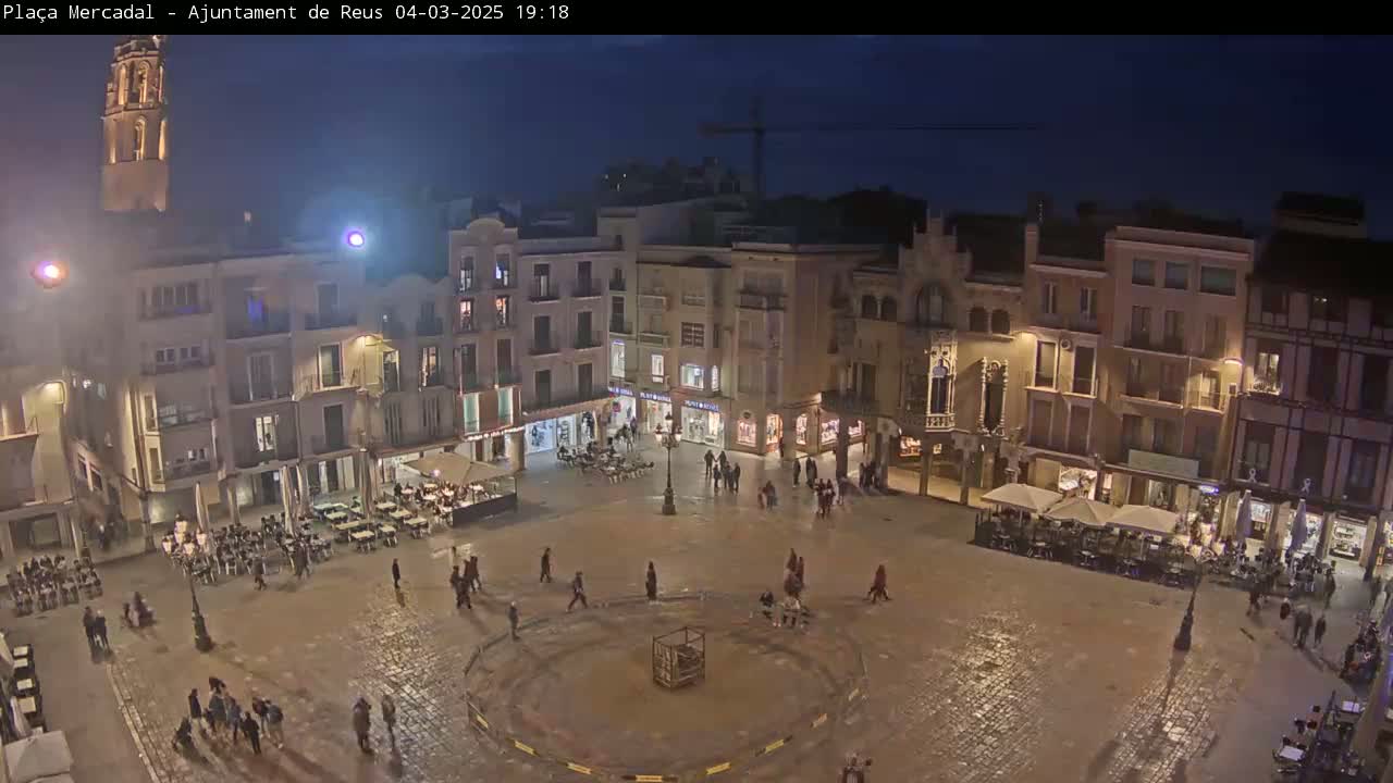 A nighttime view of a town square with several multi-story buildings surrounding it, people walking around, and outdoor seating areas at various restaurants.