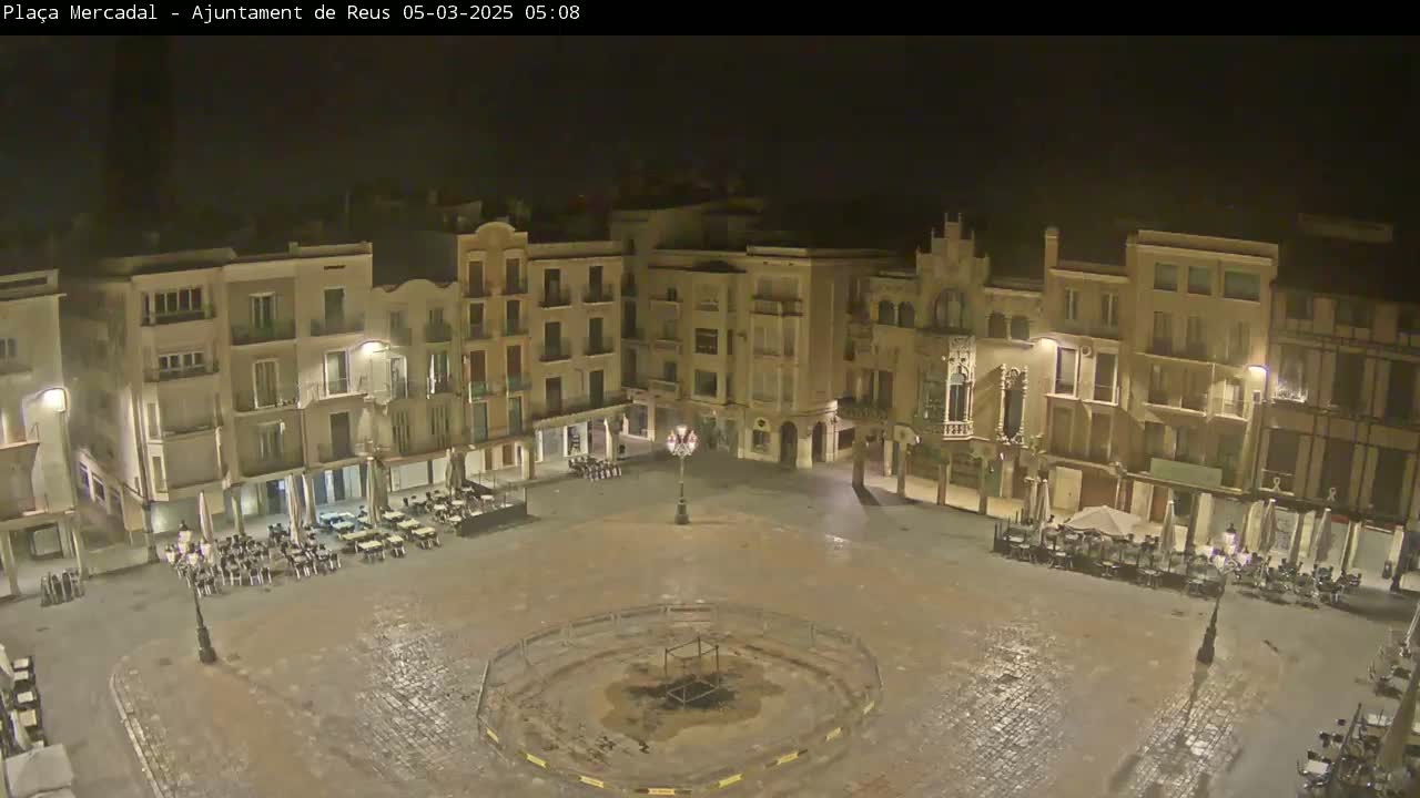 A mostly empty town square at night, lit by streetlights, with several outdoor cafe tables and chairs, appears slightly wet.