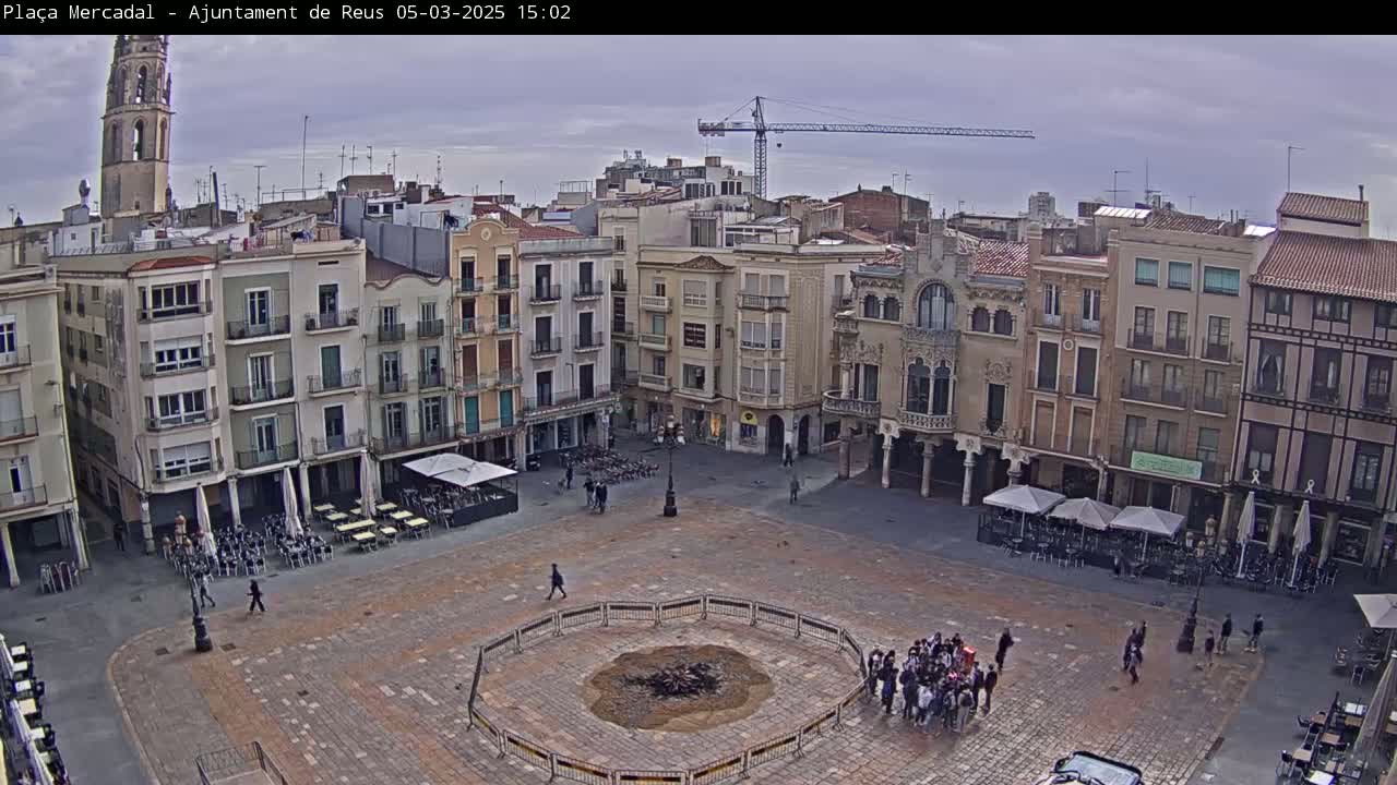 An overcast day reveals a large, paved town square surrounded by multi-story buildings, with a small group of people gathered around a central, circular area.