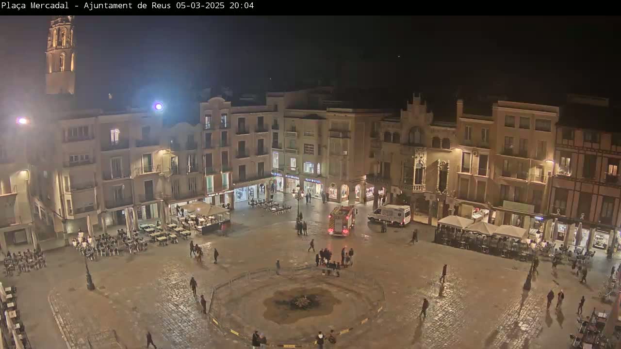 A nighttime view of a town square with several buildings surrounding it, some with outdoor seating, and a fire truck parked in the center, with a wet, possibly rain-slicked surface.