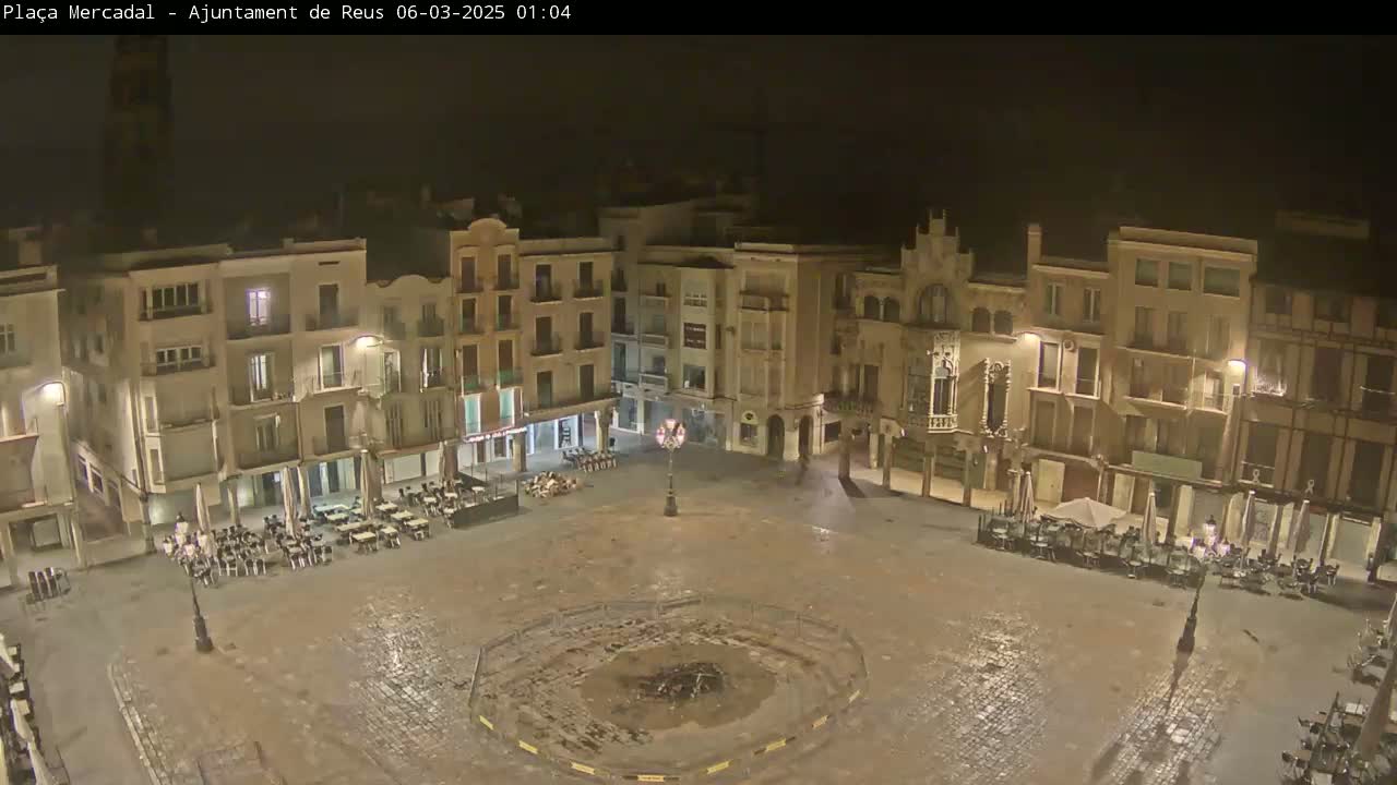A mostly empty town square at night, surrounded by multi-story buildings with outdoor seating areas, is slightly wet from recent rain.